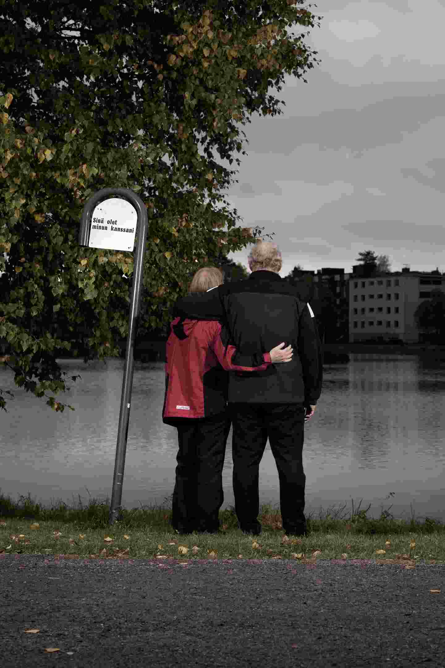 Two people stand by the pond and hug. Next to them is a sign that reads, "You are with me."