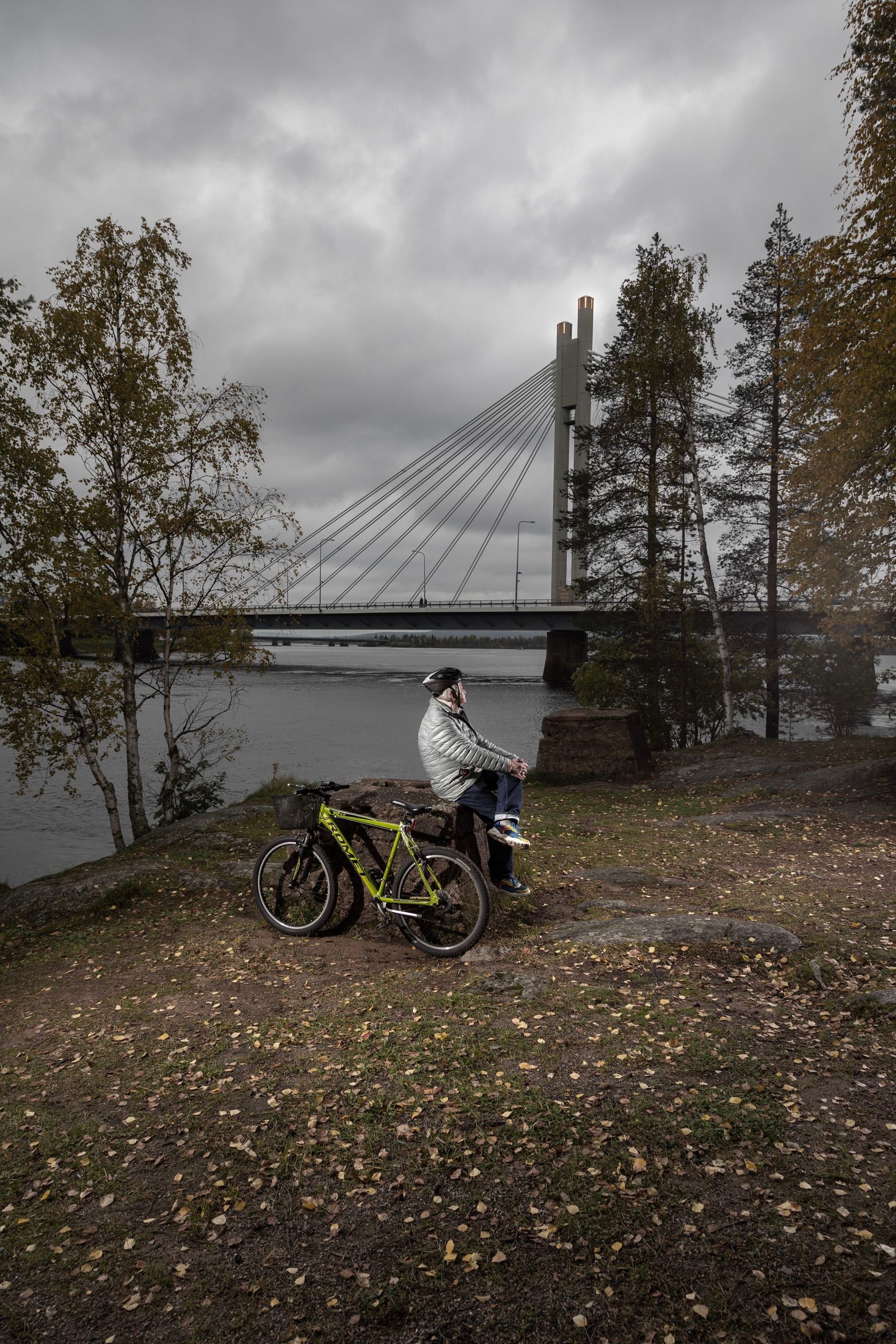 A person sits on a large stone outside, looking at the scenery. He wears a bicycle helmet and there is a parked bike next to him.