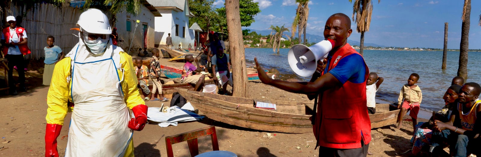 A Red Cross worker in protective gear and another person with a megaphone give instructions to people in a lakeside village.