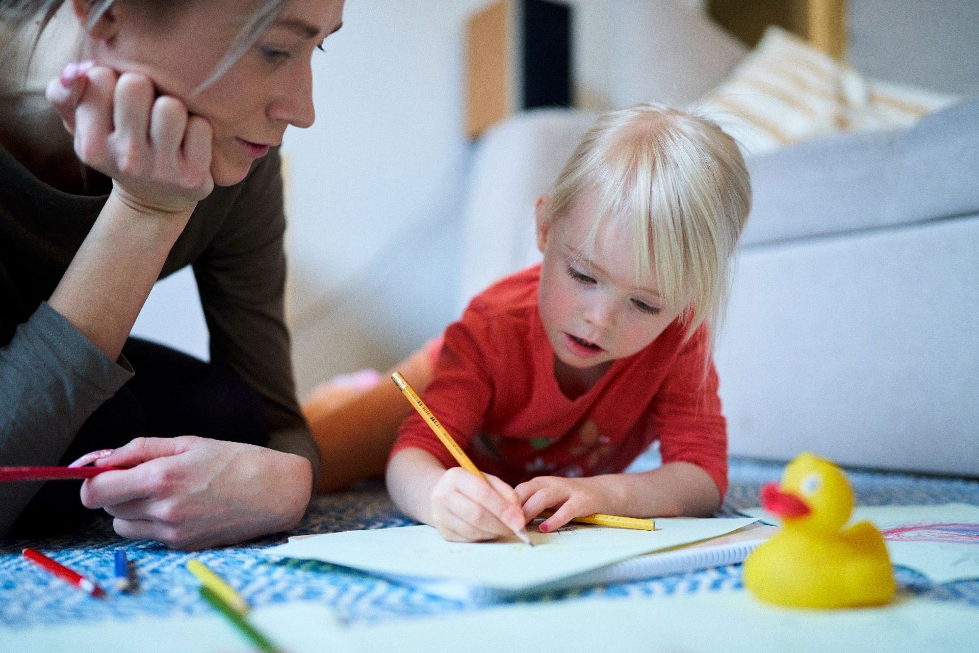 A child draws on a paper lying on the floor. An adult watches their drawing.