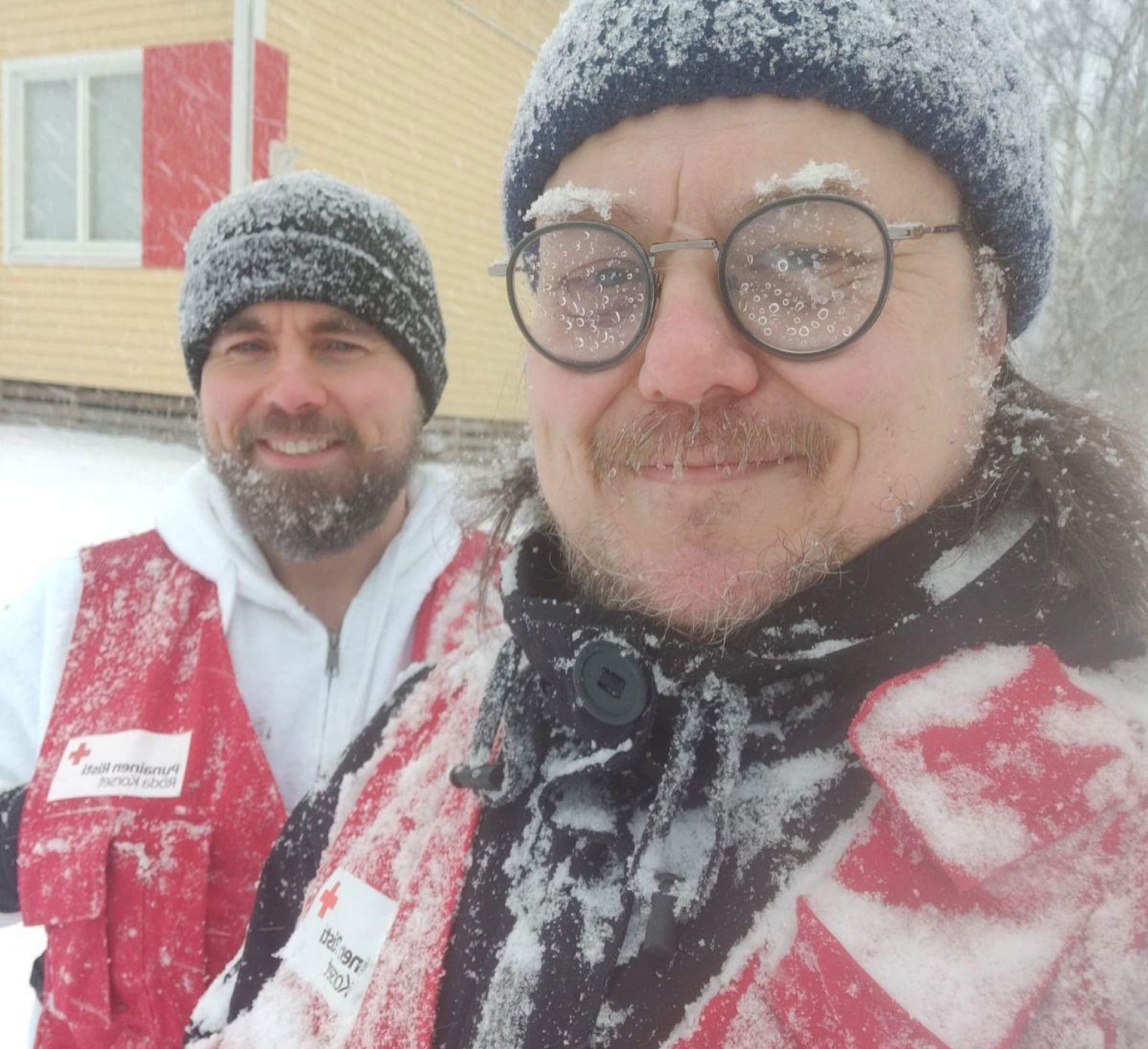 Teemu Hiilinen and Vesa Vahermaa are standing and smiling in very snowy weather, wearing Red Cross volunteer vests.