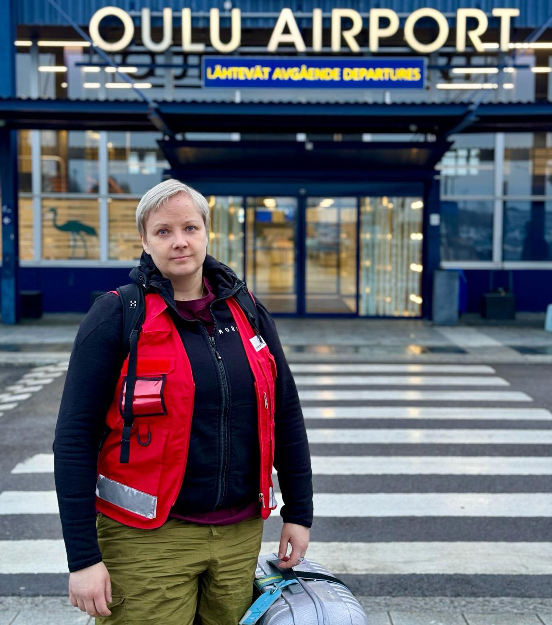 Anni Walli is standing in front of Oulu Airport in Red Cross work clothes, holding a suitcase.