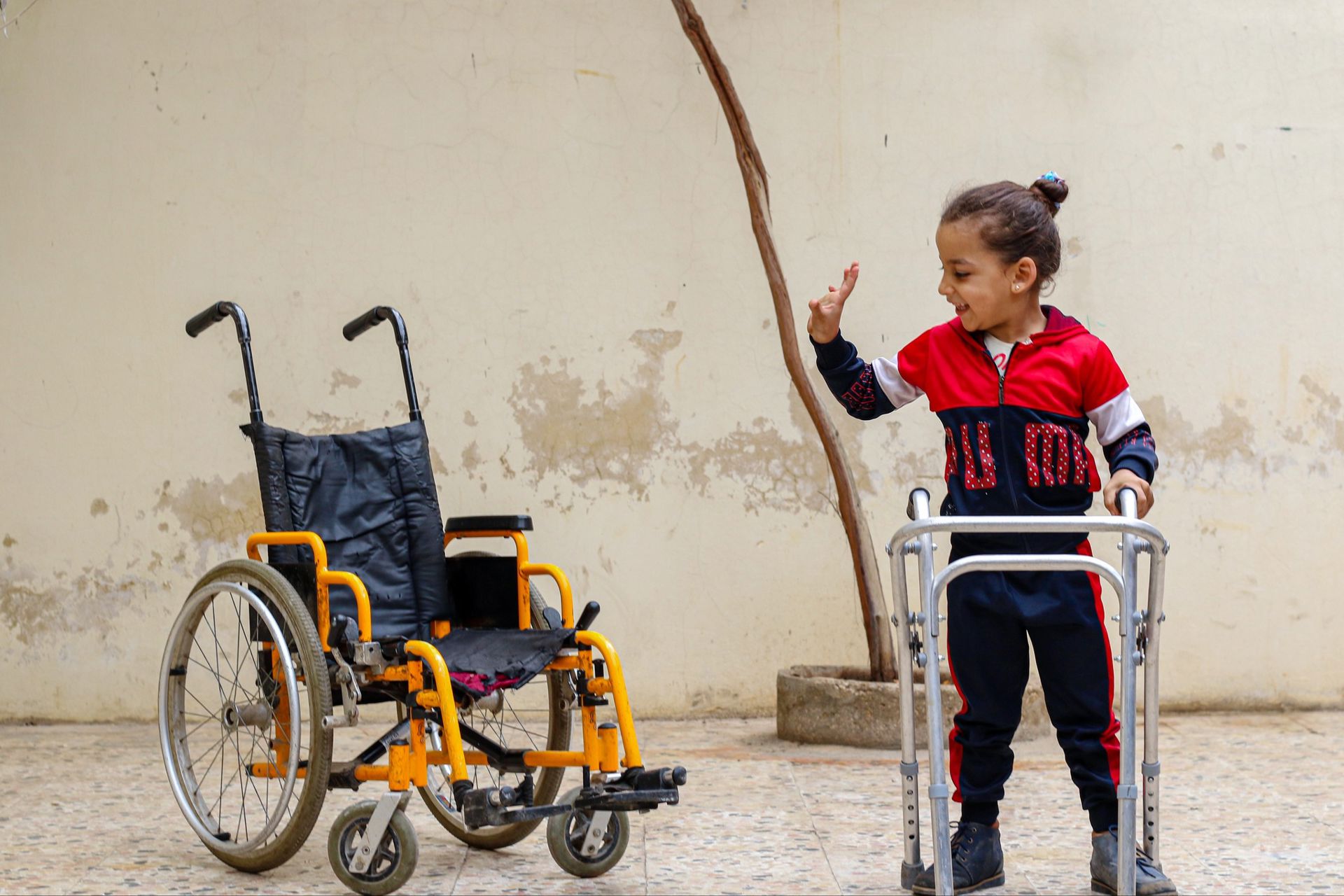 A smiling girl named Islam, standing with the help of a walker, waves at a wheelchair.