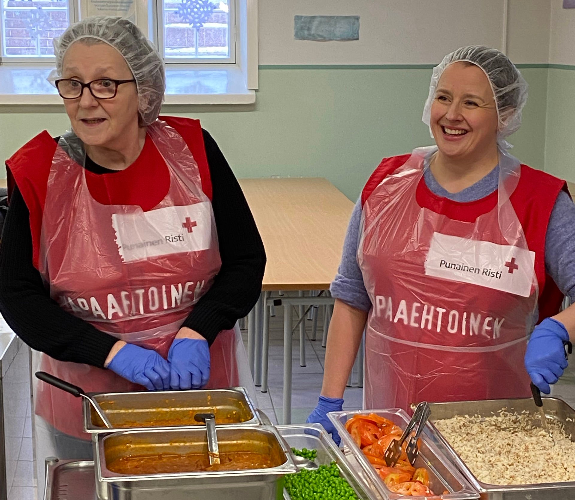 Two smiling food aid volunteers are distributing food, wearing protective caps and Red Cross volunteer vests.