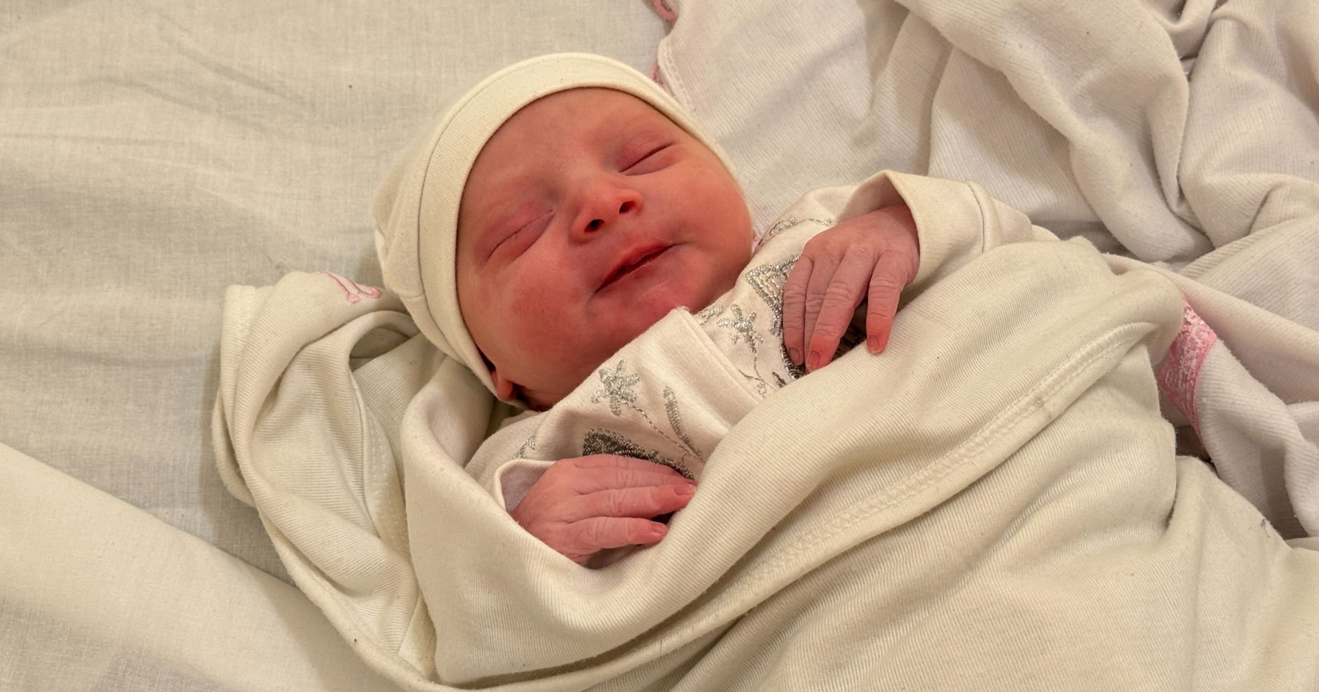 A newborn baby lies with eyes closed, wearing a white hat and a content expression on their face.