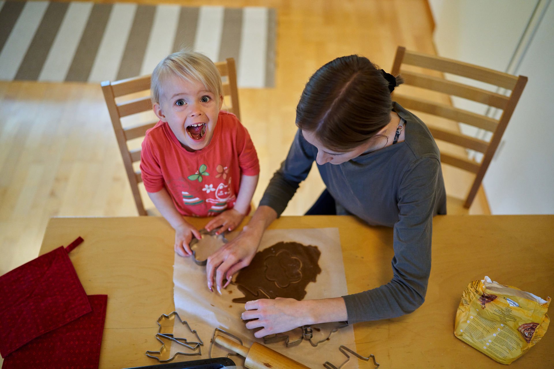 A young girl is looking at the camera with a happy expression. She is sitting at the table with her mother, baking gingerbread cookies.