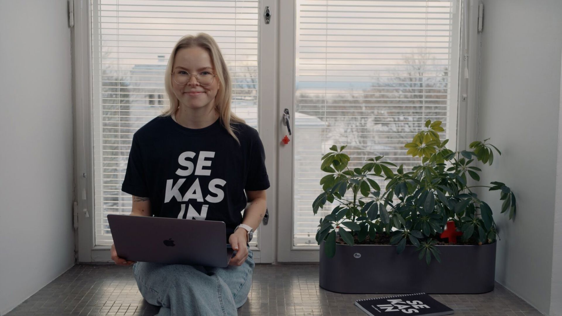 A smiling volunteer from the Sekasin chat sits in front of a window with a laptop on their lap.