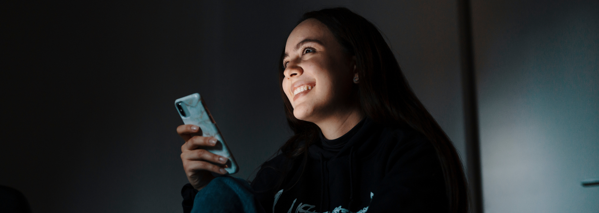 A smiling young volunteer is sitting with a phone in hand. The image is dark, but light falls on their face.