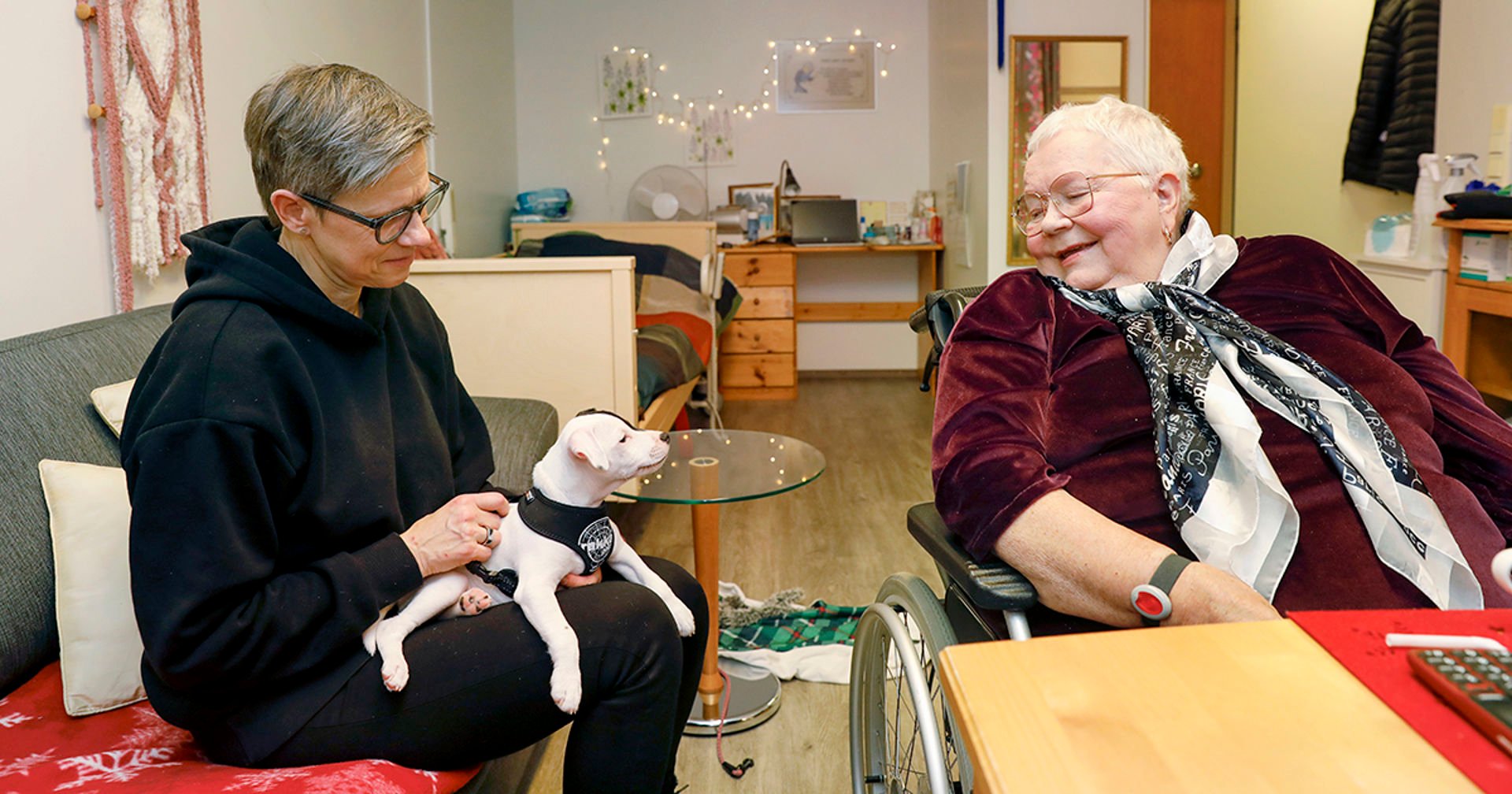 A volunteer and an older person sit around a table and smile. The volunteer has a little dog on their lap.