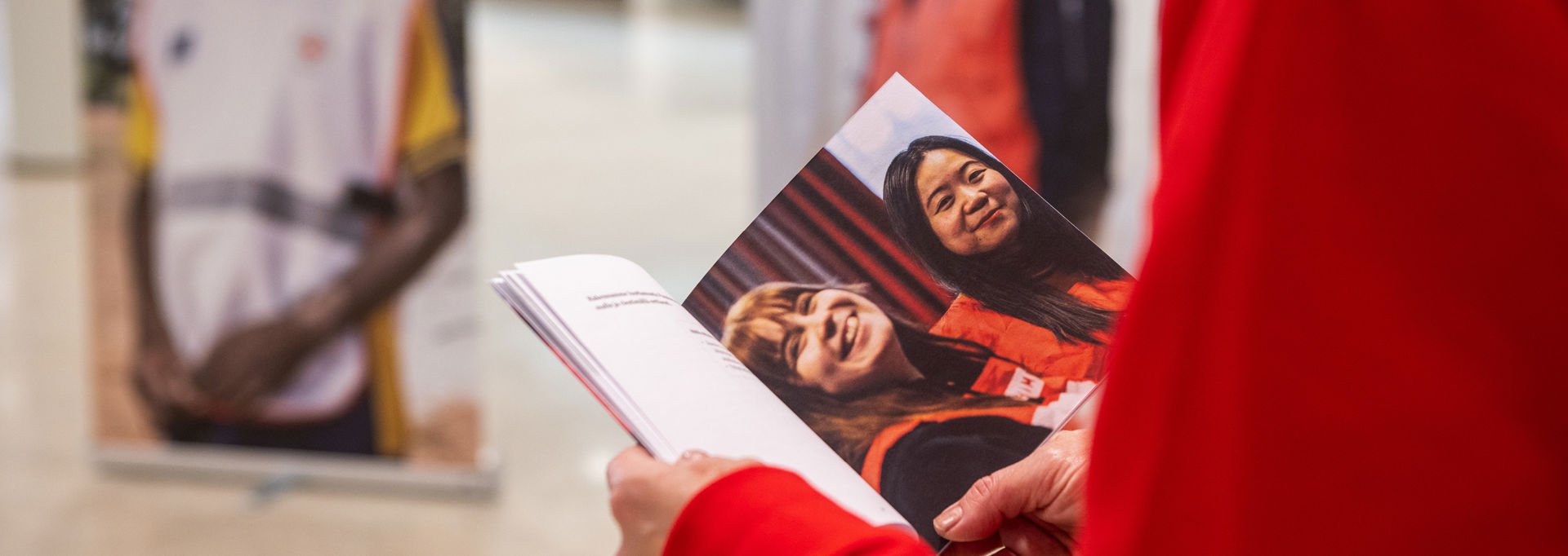 A person wearing a red shirt holds a brochure featuring a photo of two smiling individuals in Red Cross volunteer vests. In the background, blurred images of other Red Cross workers can be seen.