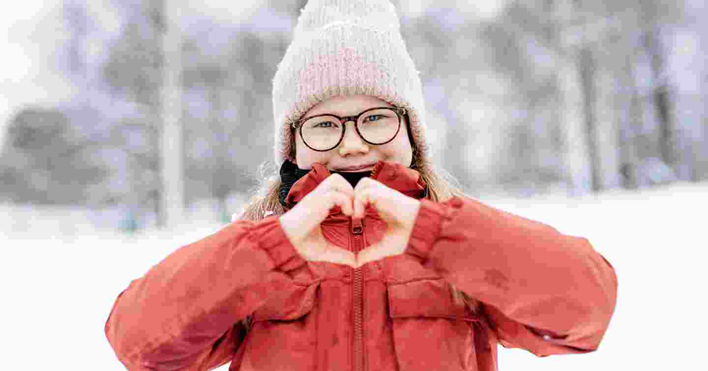 A young person smiling and making a heart sign with their hands in winter weather.