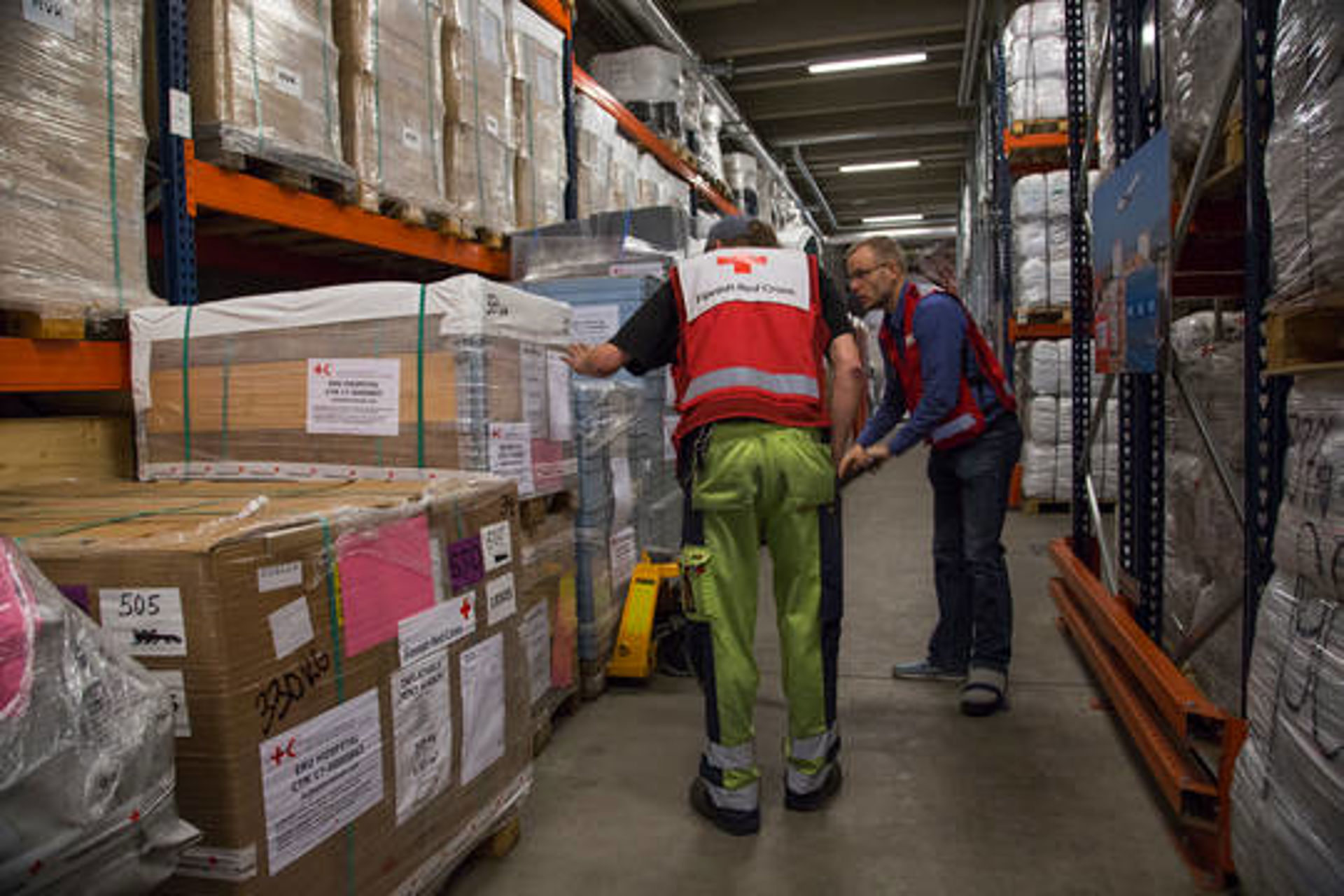 Two Red Cross employees picking up aid supplies in the Logistics Centre, which has wide, tall shelves full of supplies.