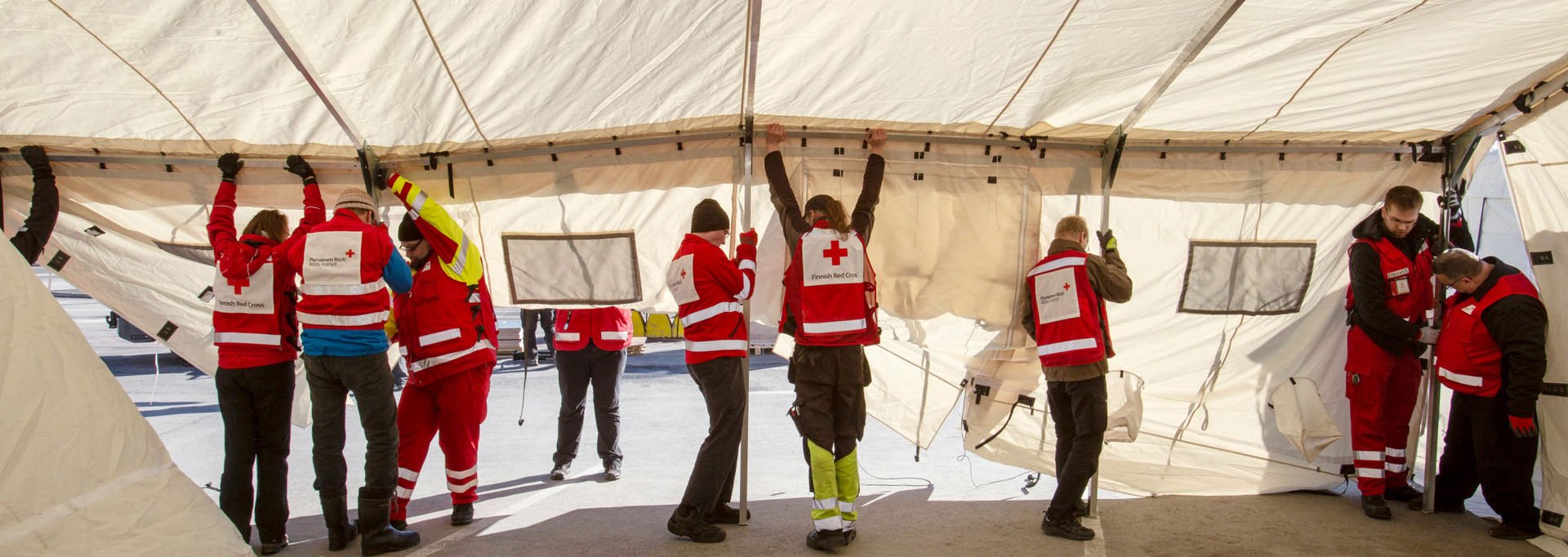 Seven people in Red Cross gear putting up a tent.