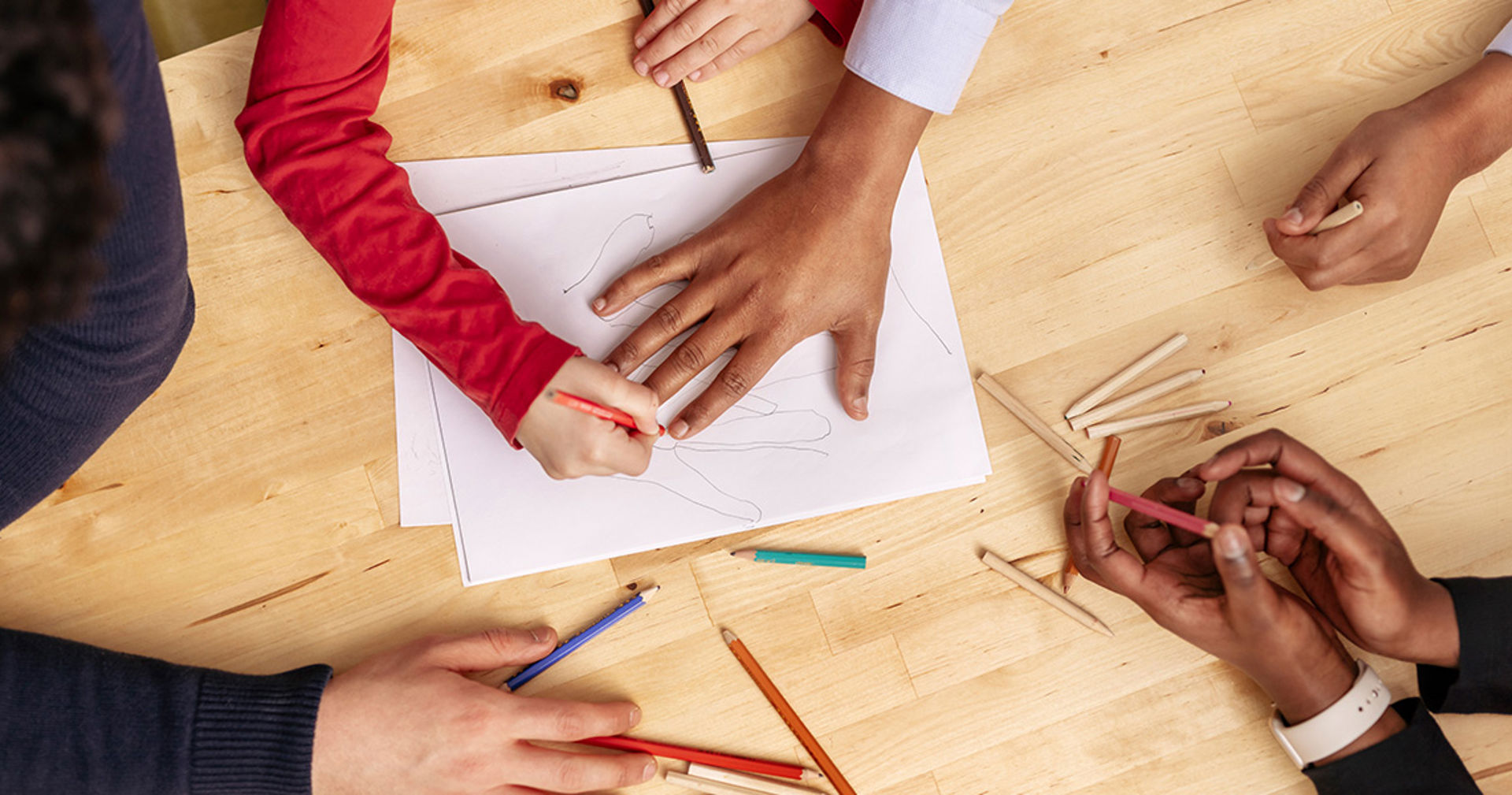 The picture shows pairs of hands drawing with wood crayons. The outline of one hand is drawn on a piece of paper on the table.