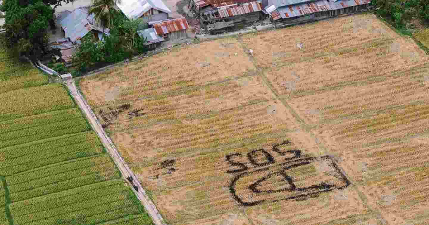 A photograph taken from above shows a field in poor condition with an SOS written on it. There are destroyed buildings at the top of the picture.
