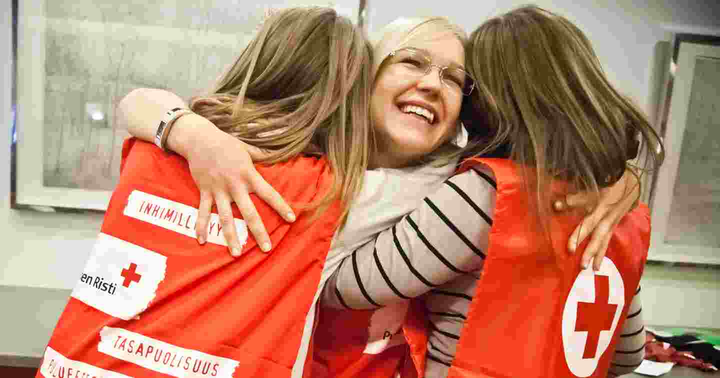 Three young women in Red Cross volunteer vests smiling and hugging each other.