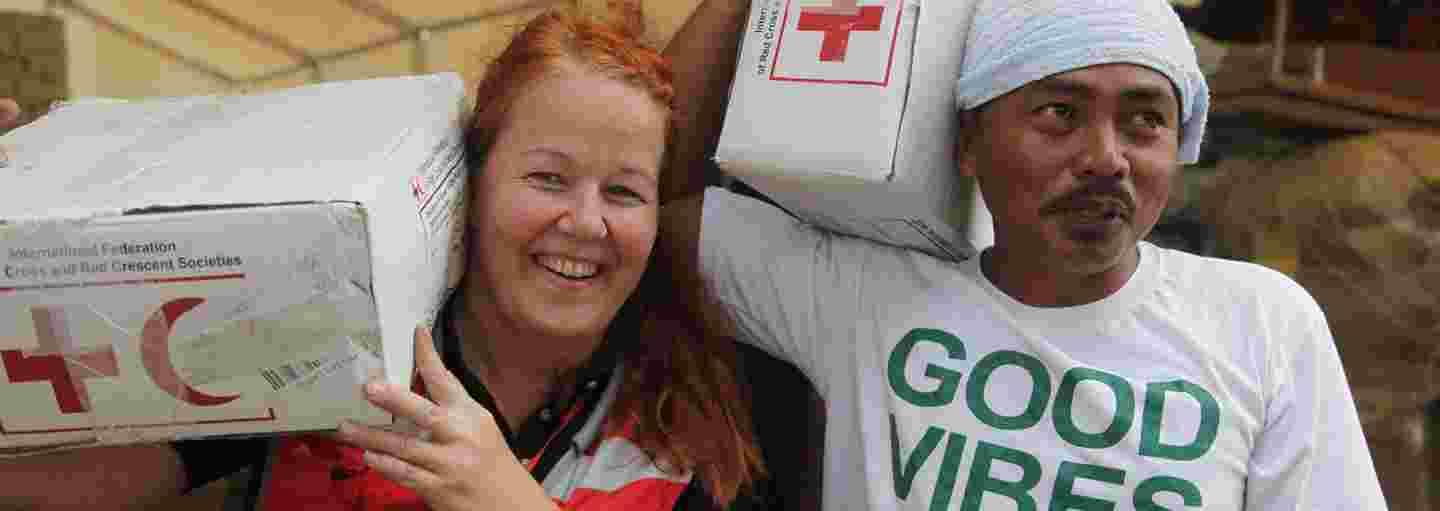 A Red Cross aid worker and a local helper carrying aid supplies with smiles on their faces.