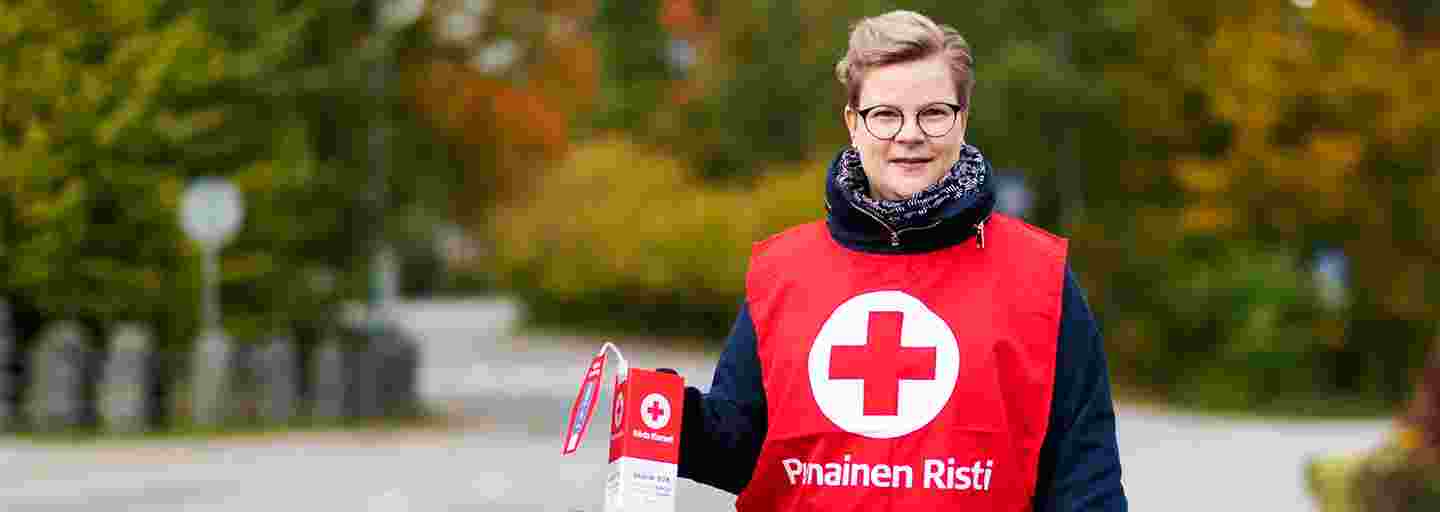 A woman dressed in the Red Cross red vest standing in an autumn view with a collection box in her hand.