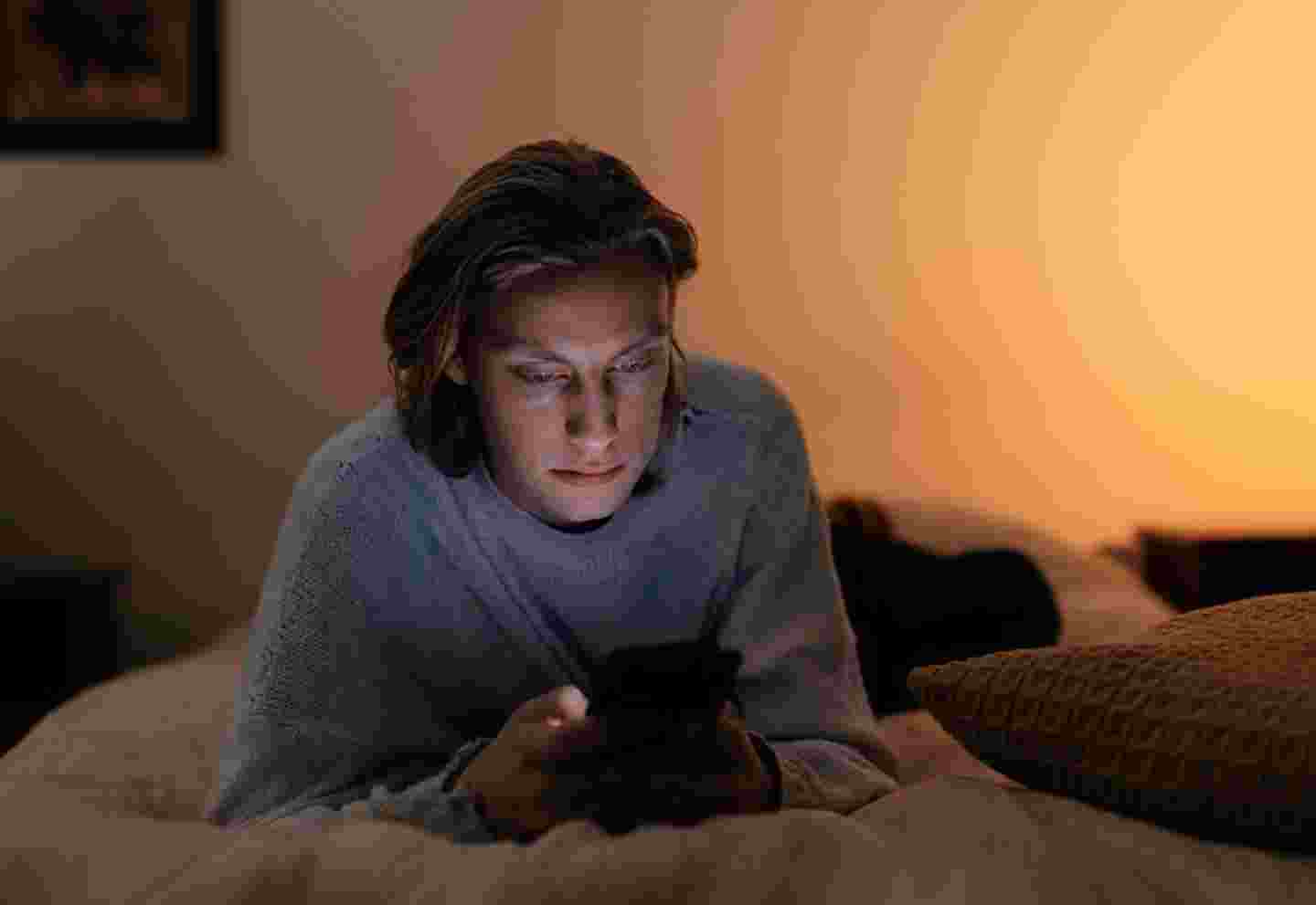 A young person using a smartphone in bed in dim lighting.